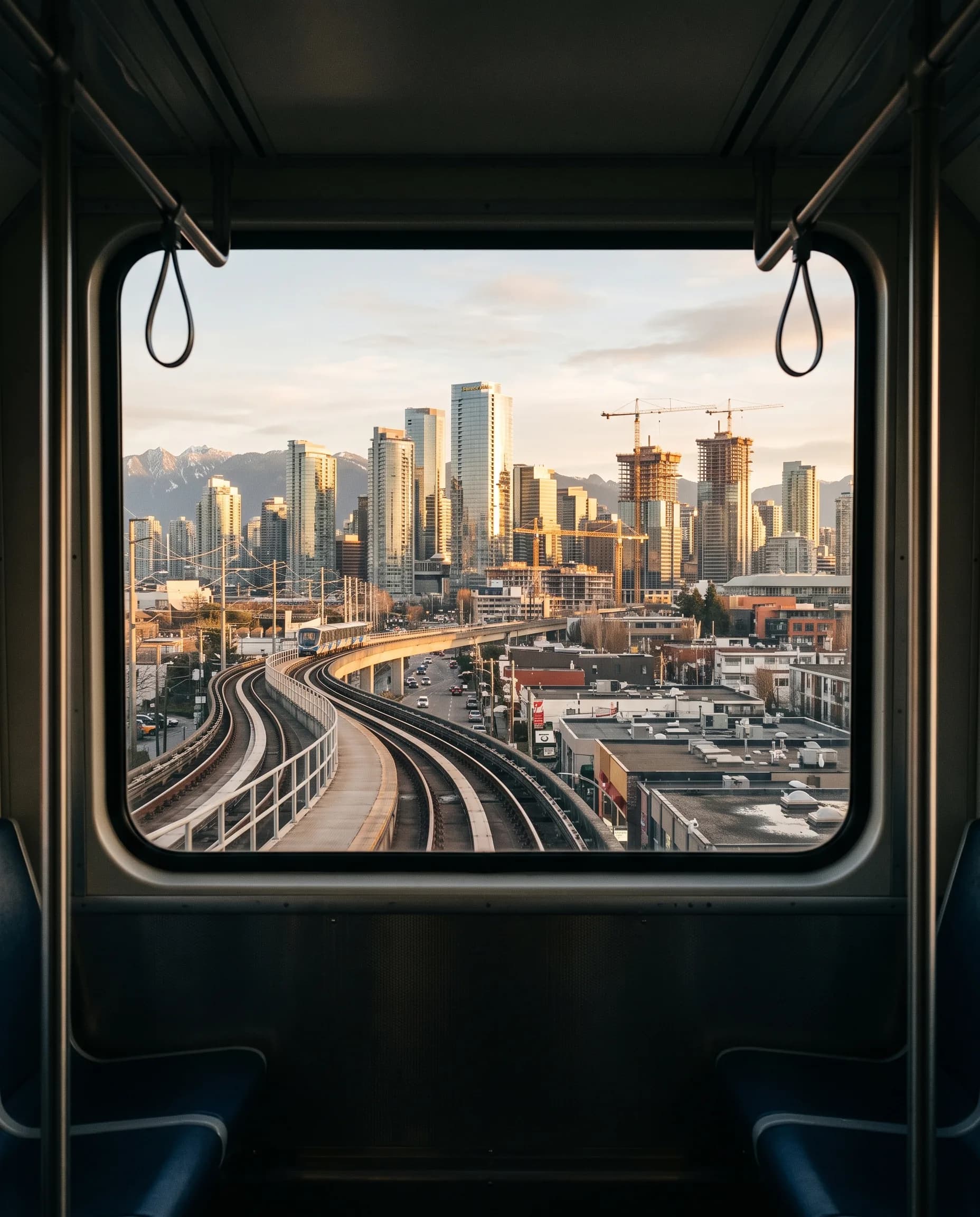 The Vancouver skyline and construction cranes at sunrise, seen through the window of an elevated SkyTrain carriage